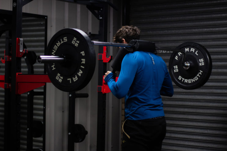 Male client with blue shirt standing with barbell on his back