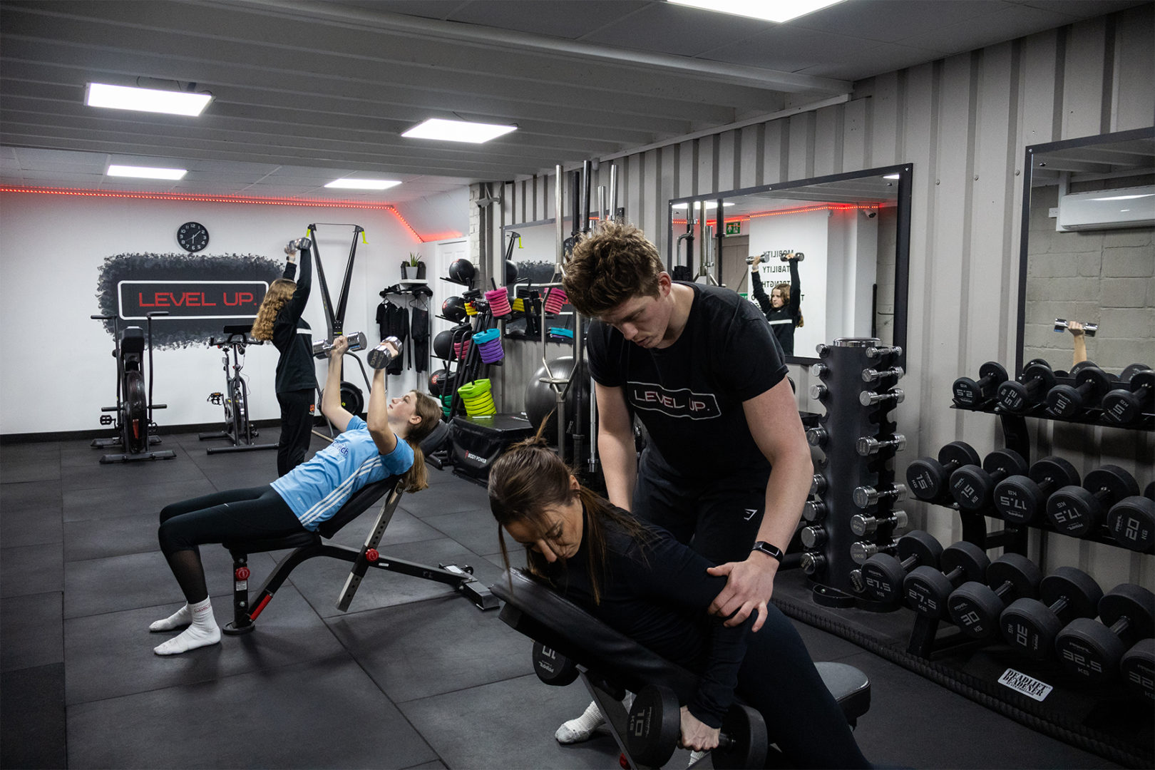 Instructor assisting client with weights during a temp class, in the background there is another client using the free weights on a bench
