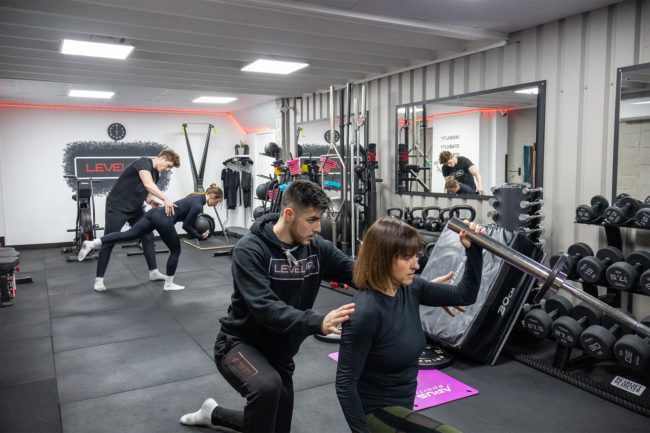 Both instructors are helping two female clients - in the background the instructor is helping the client with their form while balancing and in the foreground an instructor is helping a client with a weight