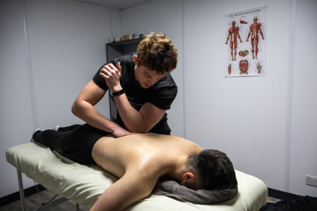 Instructor is giving a male client a back massage while the client is lying on the massage table